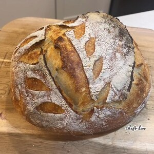 May include: A freshly baked loaf of sourdough bread with a golden-brown crust and decorative scoring. The bread is dusted with flour and sits on a wooden cutting board. The words "chefs kiss" are written on the board.