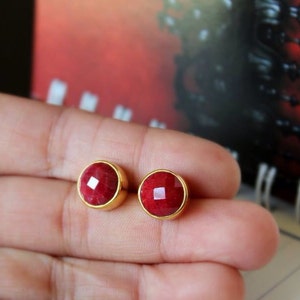 May include: A pair of gold stud earrings with red gemstone accents. The earrings are round and have a faceted design.