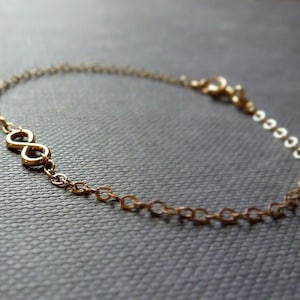 Könnte beinhalten: Ein zartes goldfarbenes Armband mit einem Unendlichkeitszeichen-Anhänger. Das Armband besteht aus kleinen, verbundenen Kettengliedern und hat einen Verschluss. Das Unendlichkeitszeichen ist ein prominentes Designelement.
