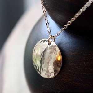 Puede incluir: Un collar de oro con un colgante redondo martillado. El colgante es de forma simple y circular, con una superficie texturizada.