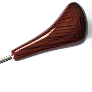 Puede incluir: Una perilla de cambio de marchas de madera con un patrón de veta de madera marrón oscuro y un eje de metal plateado. La perilla tiene una forma redondeada en forma de lágrima.