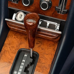 May include: A polished wooden gear shift knob positioned above a black automatic transmission gate with white lettering P, R, N, D, 3, 2, and S. The surrounding console features a glossy burl wood veneer finish with chrome accents and circular control knobs.