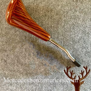 May include: A wooden gear shift knob with a brown and orange wood grain pattern and a chrome metal shaft. The shaft is bent at a 90-degree angle and ends with a black plastic tip.