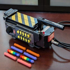 May include: A black and yellow Ghostbusters proton pack toy with a black handle and a black cord. The toy is sitting on a wooden surface with a set of rainbow colored markers.