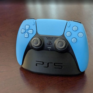 May include: A light blue and black PlayStation 5 controller on a black base. The controller has a symmetrical design with buttons and joysticks. The base has the text "PS5" printed on it.