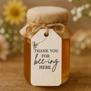 May include: A small glass jar of golden honey, with a burlap lid and twine tie. A white tag reads "THANK YOU FOR bee-ing HERE" with a bee illustration. A wedding or party favor. The jar contains approximately 200 ml of honey.