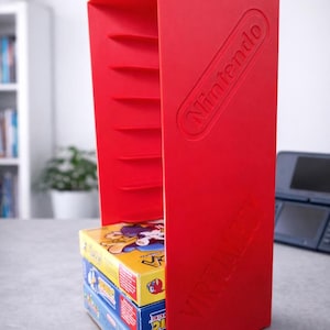 Op de afbeelding: Een felrode Nintendo-spelopslagunit met het Nintendo-logo in reliëf. De unit bevat verschillende speldozen, met de woorden "Virtual Boy" aan de zijkant gedrukt. De unit staat op een grijze ondergrond.