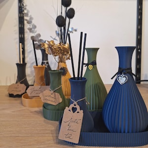 May include: An assortment of decorative vases in various colours and styles, including blue, green, gold, and black. Some vases have a ribbed texture. The vases are arranged on a wooden surface with small decorative tags.