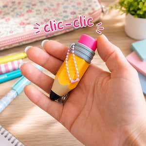Peut inclure: Un taille-crayon en forme de crayon jaune avec une gomme rose au sommet, une virole gris métallisé et une pointe noire. Il comporte une petite chaînette à billes rose sur le côté et le texte clic-clic écrit en rose au-dessus de l'objet.