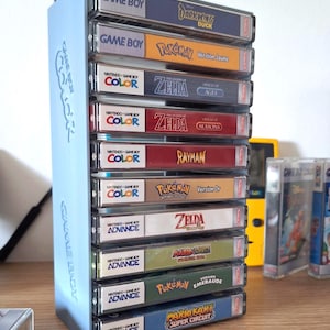 Op de afbeelding: Een lichtblauwe verticale opbergstandaard voor Game Boy-cartridges, met tien spellen waaronder Darkwing Duck, Pokémon Gele Versie, Zelda Oracle of Ages, Zelda Oracle of Seasons, Rayman, Pokémon Gouden Versie, Zelda Minish Cap, Mario & Luigi, Pokémon Smaragd en Mario Kart.