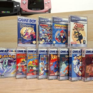 May include: A collection of 15 vintage Game Boy and Game Boy Color cartridges, including titles such as Pokemon, Zelda, and Darkwing Duck. The cartridges are arranged in rows on a wooden surface.