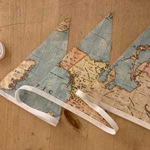 May include: A set of three blue and beige fabric pennants with a world map design. The pennants are connected with white ribbon.