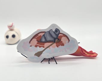May include: A whimsical, handmade clay sculpture featuring a small, white figure with a baseball cap and a cartoon-style boat with a person inside. The boat is painted with a paddle and a gray object. The sculpture is set against a white background.