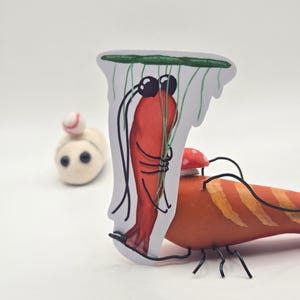 Peut inclure: Une sculpture fantaisiste de courge orange en forme de crevette, avec des pattes et des antennes en fil de fer noir. Un autocollant d'une crevette rouge aux yeux noirs est attaché. Une petite créature feutrée blanche avec une tête en forme de balle de baseball est en arrière-plan.