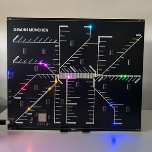 Könnte beinhalten: Eine schwarze, beleuchtete Karte des S-Bahn München-Netzes. Die Karte zeigt weiße Linien, die Routen und Stationsnamen darstellen. Farbige LED-Leuchten heben verschiedene Stationen hervor. Der Text "S-BAHN MÜNCHEN" befindet sich oben.