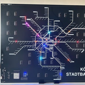 Könnte beinhalten: Eine schwarze, beleuchtete Karte des öffentlichen Nahverkehrsnetzes der Kölner Stadtbahn. Die Karte zeigt ein Netz von Linien mit verschiedenen farbigen Lichtern und den weißen Text "KÖLN STADTBANH". Die Karte ist mit einem USB-Kabel verbunden.