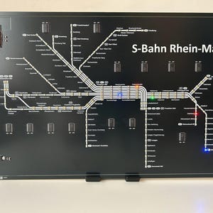 Könnte beinhalten: Schwarzes elektronisches Display mit einer detaillierten Karte des S-Bahn Rhein-Main-Bahnnetzes. Die Karte enthält Stationsnamen und Linien, mit beleuchteten Abschnitten in verschiedenen Farben. Der Text "S-Bahn Rhein-Main" ist deutlich sichtbar.