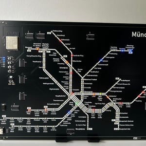Könnte beinhalten: Schwarzes elektronisches Display mit einer detaillierten Karte des Münchner U-Bahn-Systems. Die Karte ist mit farbigen Lichtern beleuchtet, die verschiedene Linien und Stationen hervorheben. Das Wort "München" wird oben angezeigt.