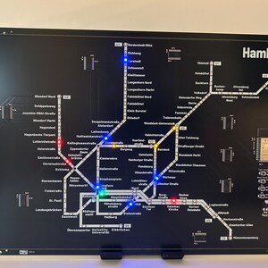 Könnte beinhalten: Schwarz beleuchtete Karte des Hamburger U-Bahn-Systems. Die Karte zeigt weiße Linien und Stationsnamen, mit farbigen LED-Leuchten, die Routen anzeigen. Das Wort "Hamburg" ist in weißer Schrift oben rechts dargestellt.