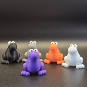 May include: Five colorful frog figurines in black, gray, orange, purple, and white. Each frog has large, white eyes and a rounded body with four small feet. The frogs are arranged on a dark surface.
