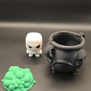 May include: A white mummy figurine, a black cauldron, and a green, cloud-shaped object are arranged on a dark surface. The cauldron has handles and a skull design. The mummy has black eyes. The cloud-shaped object has a flat base.