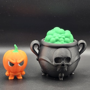 May include: An orange pumpkin figure with an angry face stands next to a black cauldron. The cauldron has a skull design and is filled with green foam. The cauldron has two handles and three feet.