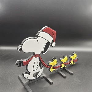 May include: A Snoopy Christmas decoration featuring Snoopy wearing a Santa hat and scarf, pulling three Woodstock figures. The figures are in red, white, black, and yellow. The decoration is mounted on a black base.