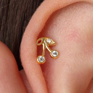 May include: A gold-colored cherry-shaped earring with three clear, round gemstones. The earring is worn on the ear cartilage. The cherry design includes two round gems for the cherries and one for the leaf.