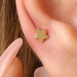 May include: A close-up of a gold-coloured star-shaped earring. The earring is a small, five-pointed star, and it is positioned on the ear. The skin around the ear is visible, and the earring appears to be a stud. The earring is a simple design.