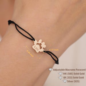 Könnte beinhalten: Ein goldfarbenes Armband mit einem Pfotenabdruck-Anhänger mit dem eingravierten Namen "Leo". Der Anhänger ist mit einem schwarzen, verstellbaren Makramee-Paracord verbunden. Der Anhänger misst 1,19 cm x 1,17 cm. Das Armband besteht aus 14K (585) Massivgold, 8K (333) Massivgold oder Silber (925).