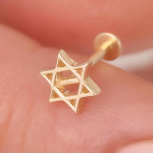 Pode incluir: Um brinco dourado com um design de estrela de seis pontas. A peça de joalharia possui um pino fino e um fecho circular plano, exibindo uma forma geométrica de estrela com estrutura aberta e um aspeto polido e metálico.