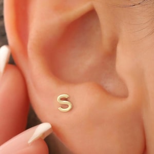Pode incluir: Um brinco de letra "S" dourada em uma orelha. O brinco é pequeno e simples, com um acabamento polido. A orelha está em foco, com o brinco centrado na imagem.