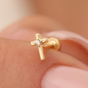 Może przedstawiać: Złoty kolczyk typu sztyft z małym motywem krzyżyka i pojedynczym, przezroczystym, okrągłym kamieniem osadzonym pośrodku. Kolczyk posiada płaskie zapięcie oraz gładkie, polerowane wykończenie, co nadaje mu prosty i przejrzysty wygląd.