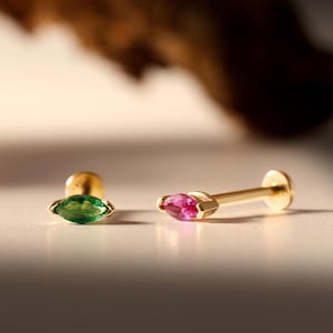 Könnte beinhalten: Zwei goldene Ohrstecker mit flacher Rückseite und marquise-geschliffenen Edelsteinen. Ein Ohrstecker ist mit einem leuchtend grünen Stein besetzt, der andere mit einem leuchtend pinken Stein. Beide haben einen goldenen Stift und einen runden Verschluss.
