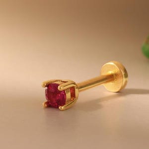 May include: A gold-coloured labret stud with a vibrant, round, red gemstone. The stud features a post and a flat disc backing. The gemstone is held in place by four prongs. The jewellery is designed for body piercing.