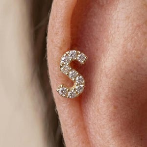 May include: A gold-colored stud earring in the shape of the letter "S" is shown. The letter is encrusted with small, sparkling, clear gemstones. The earring is set against the backdrop of a person's ear.