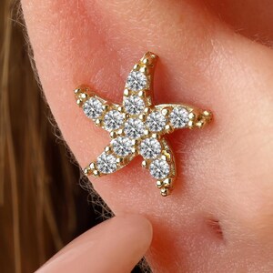 May include: A gold starfish-shaped earring stud featuring a textured metallic surface and multiple small, round, clear, faceted stones embedded along the five arms and center of the design, creating a sparkling appearance on the earlobe.