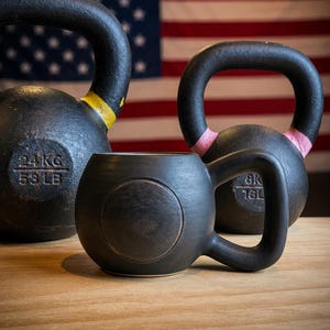 May include: A black ceramic mug shaped like a kettlebell with a thick handle, positioned in front of two iron kettlebells. The left kettlebell is marked 24KG and 53 LB, while the right kettlebell is marked 8KG and 16 LB, with a blurred American flag behind.