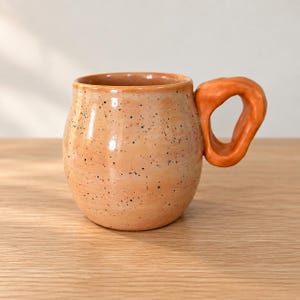 May include: A round ceramic mug with a speckled light orange glaze and a thick, solid orange handle. The mug features a smooth, rounded body with a wide opening and a distinct, irregular handle shape attached to the side of the vessel.