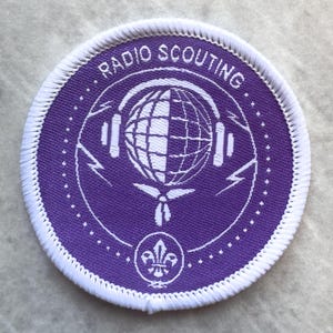 May include: A circular embroidered patch with a purple background and white border. It features a globe wearing headphones, a neckerchief, and a fleur-de-lis emblem at the bottom. The text RADIO SCOUTING is arched across the top in white lettering.