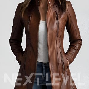 Puede incluir: Chaqueta de cuero marrón con cuello alzado y corte entallado. La chaqueta presenta cierre de cremallera, bolsillos y diseño largo. El texto "NEXT LEVEL" es visible en la parte inferior de la chaqueta.