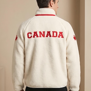 Può includere: Una giacca in pile color bianco sporco con colletto alto e finiture rosse. Sul retro è presente la scritta CANADA in lettere rosse in grassetto, con un emblema a foglia d'acero rosso ricamato su ogni manica. Giacca a maniche lunghe in tessuto felpato.