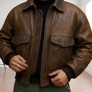 Könnte beinhalten: Braune Leder-Bomberjacke mit Reißverschluss, zwei Vordertaschen und geripptem Kragen und Bündchen. Die Jacke hat einen Vintage-Look. Die Person trägt einen schwarzen Rollkragenpullover.