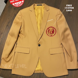 Puede incluir: Un blazer color beige con cierre de un botón, un bolsillo en el pecho y dos bolsillos delanteros. El blazer tiene un forro dorado y un emblema rojo y dorado en el pecho. El texto "FREE CUSTOM LOGO" es visible.