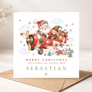 May include: A square Christmas greeting card featuring a watercolour illustration of Santa Claus and an elf flying a red and white propeller aeroplane. The text reads MERRY CHRISTMAS TO A SPECIAL LITTLE BOY SEBASTIAN, with stars and clouds.