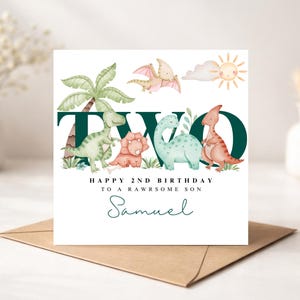 May include: A birthday card with a dinosaur theme. The card features the word "TWO" in dark green, with illustrations of dinosaurs, a palm tree, and a sun. The text reads "HAPPY 2ND BIRTHDAY TO A RAWSOME SON" and "Samuel".