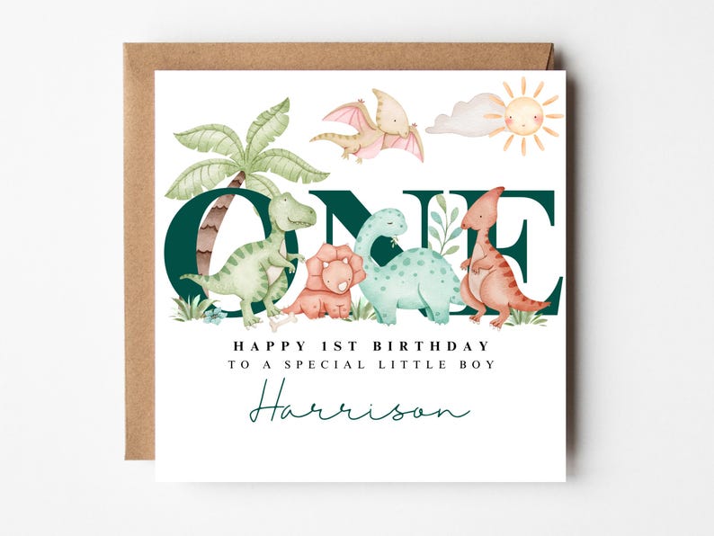 May include: A birthday card with a brown envelope. The card features watercolour illustrations of dinosaurs, a palm tree, and a sun. Text reads "ONE", "Happy 1st Birthday to a special little boy" and a name.