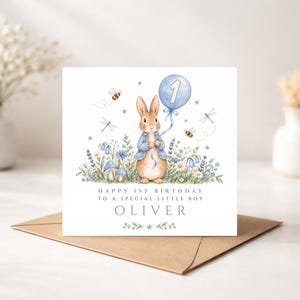 May include: A square greeting card featuring a watercolour illustration of a brown rabbit in a blue jacket holding a blue balloon with the number 1. The card includes text reading HAPPY 1ST BIRTHDAY TO A SPECIAL LITTLE BOY and the name OLIVER.