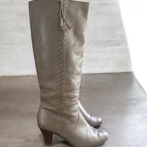 May include: A pair of knee-high taupe leather boots featuring a vertical stitched detail along the outer side, a small decorative tassel at the top, a rounded toe, and a dark brown block heel. The boots have a smooth finish and a structured shaft.