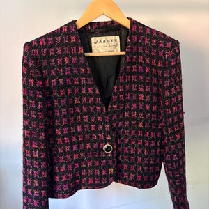 May include: A black blazer with a textured, woven pattern in vibrant pink and orange. The jacket features a single front button closure, long sleeves with button cuffs, and a black interior lining. A label inside reads JAEGER London Paris New York.